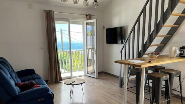 Un salon moderne avec un canapé bleu, une petite table ronde et une table à manger en bois, accompagnée de deux tabourets. Un escalier avec une rampe noire mène à un balcon d’où l’on peut admirer la vue sur la mer.