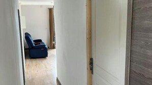 Un couloir étroit, avec une porte en bois sur la droite, qui mène à une pièce où se trouvent une chaise bleue et une valise posée sur le sol.