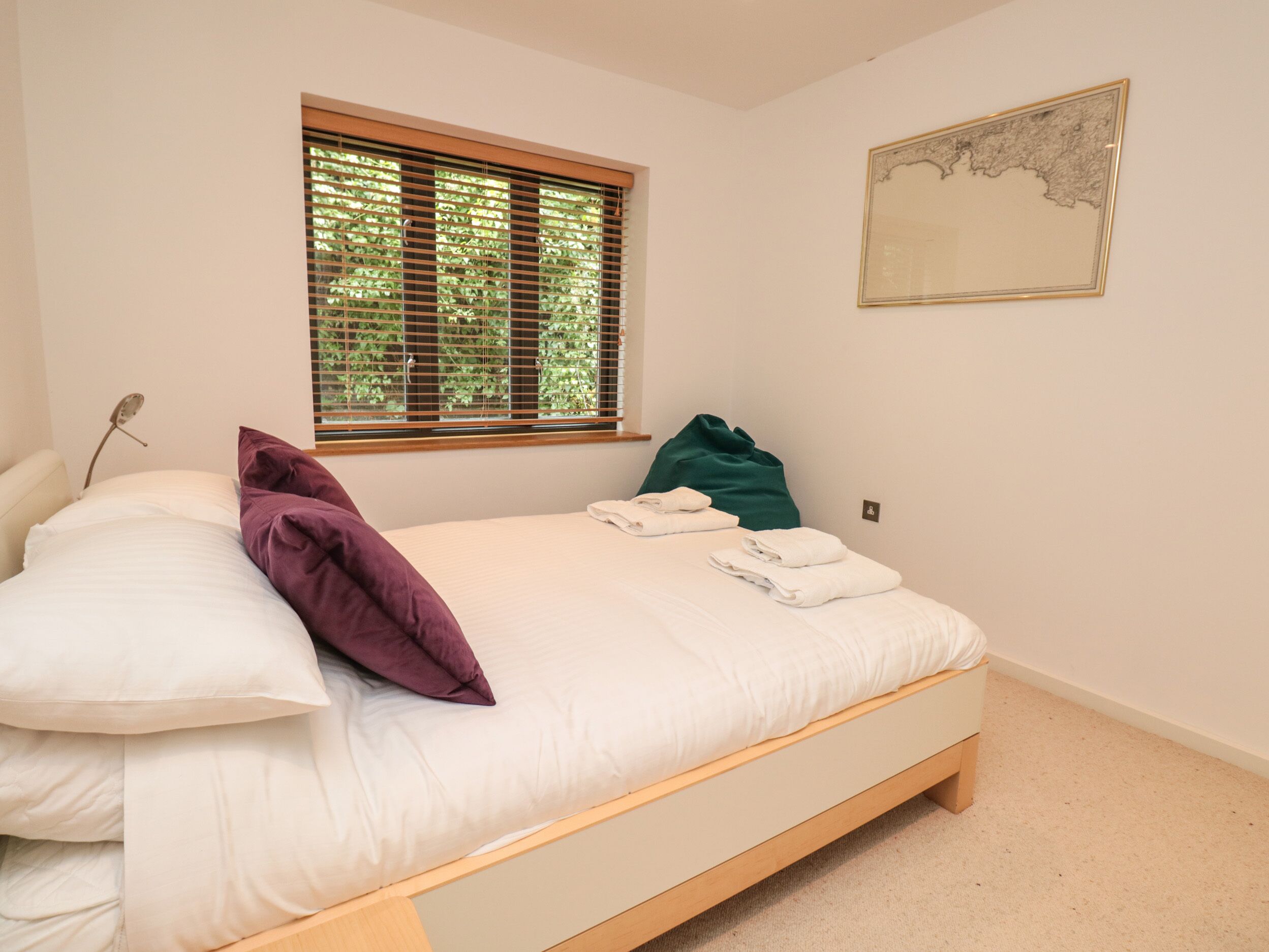 A neatly made bed with white and purple pillows, a green bag, and folded towels on a wooden bed frame in a room with a window and a framed map on the wall.