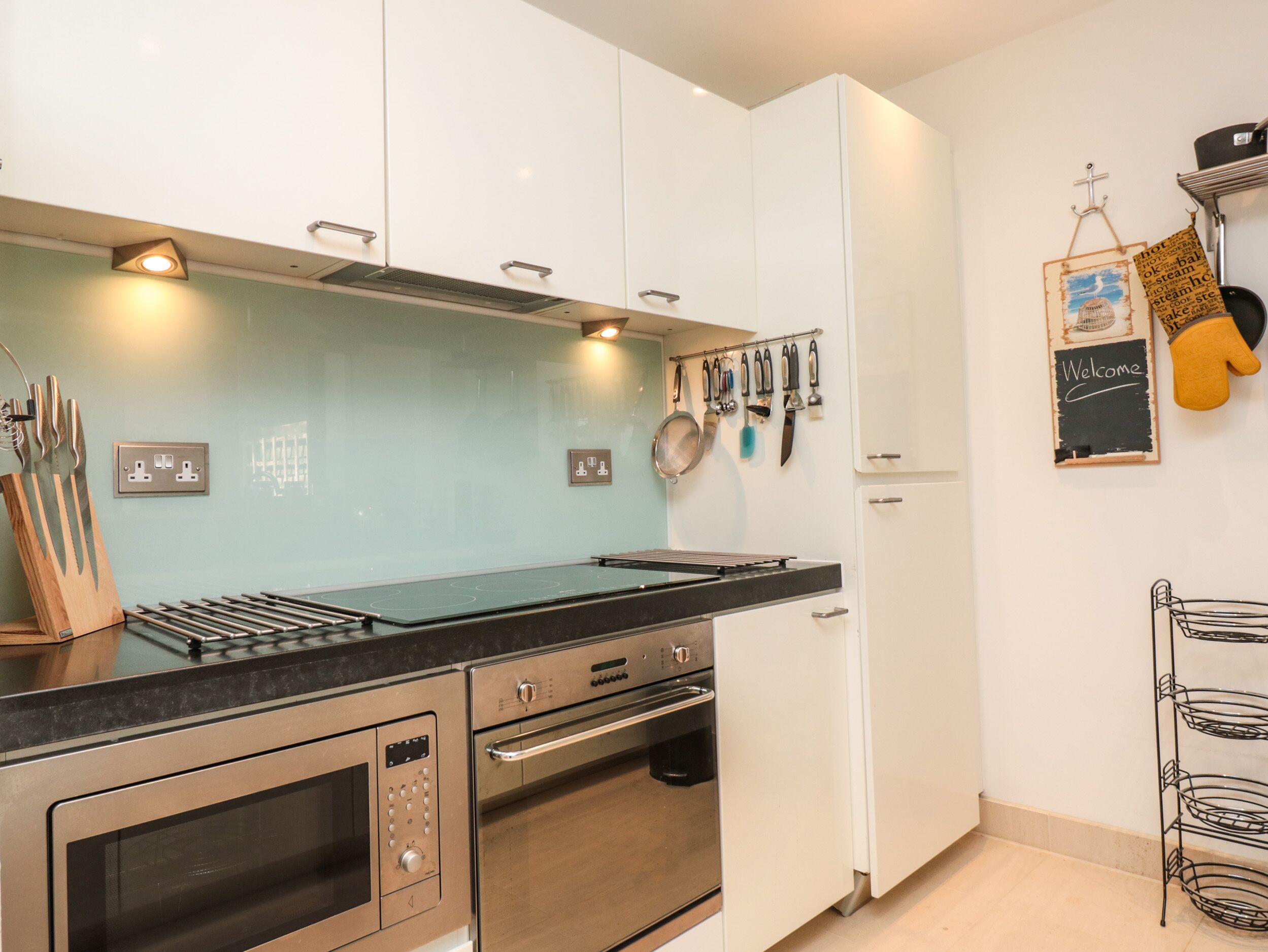 A modern kitchen with stainless steel appliances, a black countertop, and white cabinetry. There are utensils hanging on the wall and a blackboard with 'Welcome' written on it.