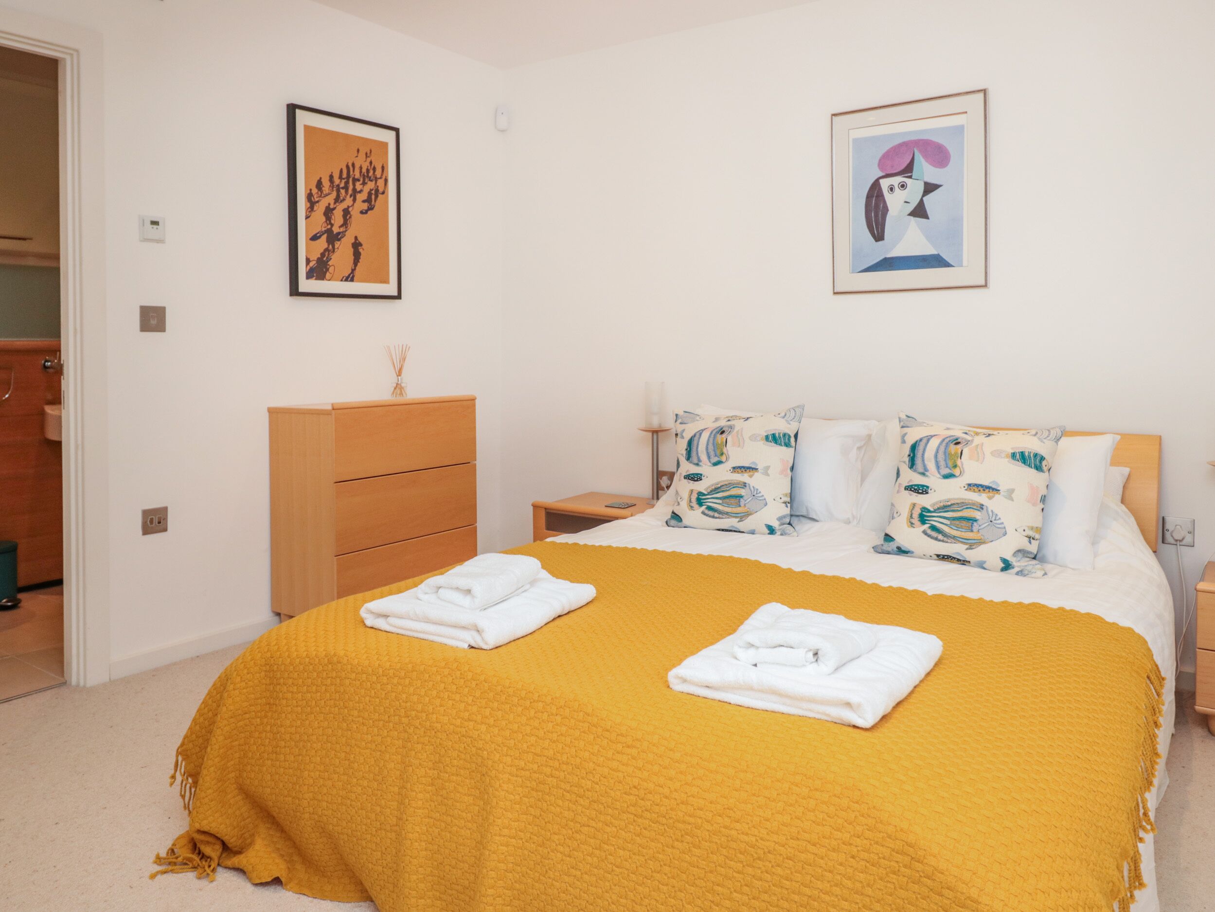 Une chambre à coucher comprenant un lit, une commode en bois, deux œuvres d’art encadrées et une table de chevet avec une lampe.