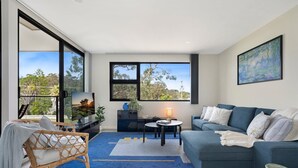 A modern living room with a large window, a blue sofa, a wicker chair, and a small table with a lamp. - Husky Sol Coastal escape, walk to beach and cafes (Huskisson)