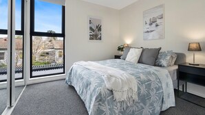 A bedroom with a bed, a nightstand, a lamp, and a large window with a view of buildings and trees. - Husky Sol Coastal escape, walk to beach and cafes (Huskisson)