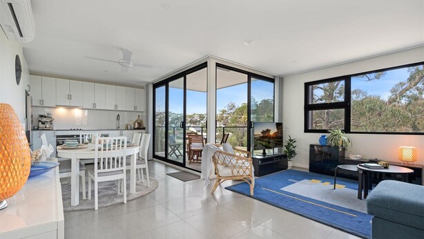 A modern kitchen with white cabinets, a dining area with white chairs, and a large sliding glass door leading to a balcony with a view of trees. - Husky Sol Coastal escape, walk to beach and cafes (Huskisson)