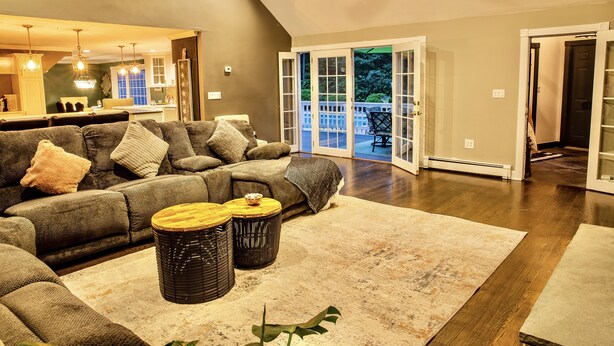 A spacious living room with a large sectional sofa, a wooden coffee table, and a large rug. There is a sliding glass door leading to a balcony with a view of trees.