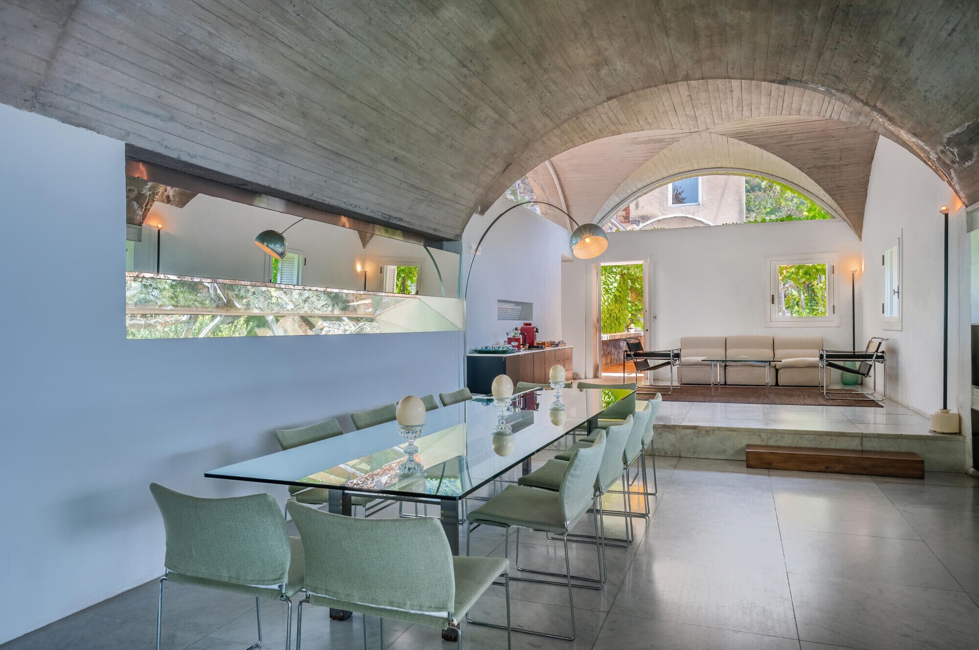 A modern interior with a glass dining table, green chairs, a sofa, and a kitchen area.