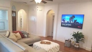 A living room with a beige sectional sofa, a white ottoman, a ceiling fan, a TV on the wall, and a potted plant. - Lovely Heights Cottage 🌅🌄 (Fenced Yard, Garage) (Houston)