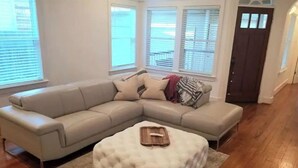 A living room with a beige sectional sofa, a white ottoman, and a wooden tray on the ottoman. - Lovely Heights Cottage 🌅🌄 (Fenced Yard, Garage) (Houston)
