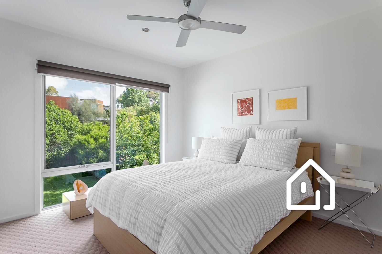 A bedroom with a large window, a bed, a ceiling fan, and two wall-mounted lamps.
