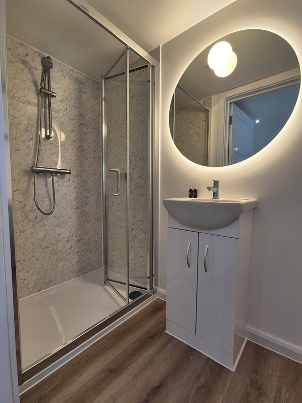 Un baño moderno con ducha de vidrio, tocador blanco con un espejo redondo y suelo de madera.