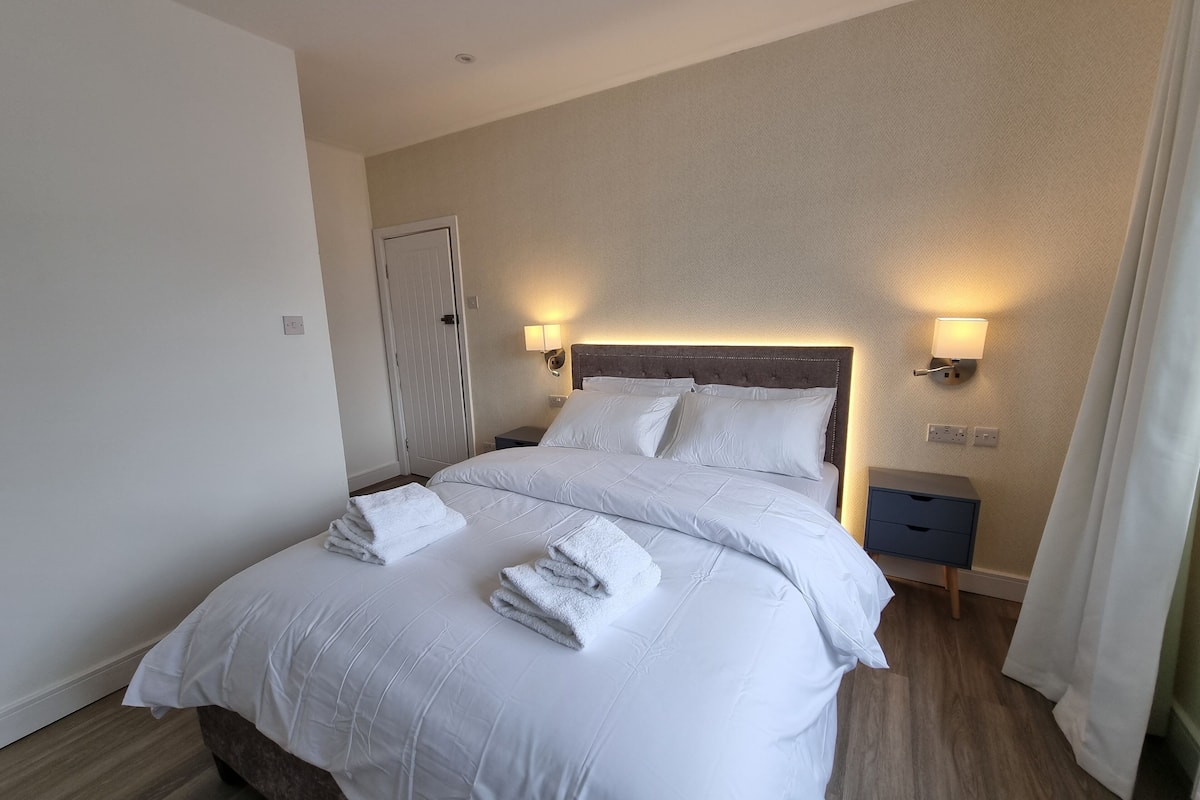 A neatly made bed with white linens and towels, a wooden headboard, two bedside lamps, and a nightstand with a drawer.
