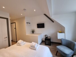 Una habitación de hotel moderna con una cama grande, un área de descanso con una silla, una mesita con un hervidor y un televisor montado en la pared.