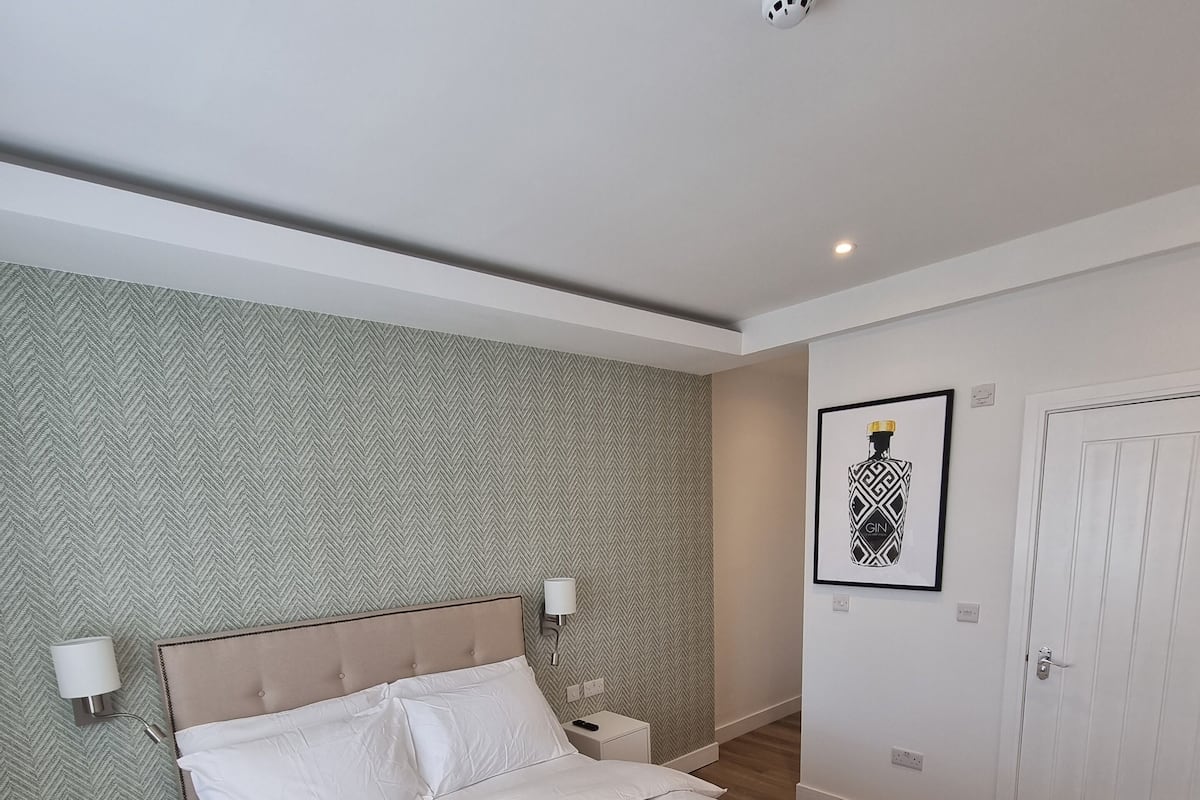 A neatly made bed with white linens and towels, a headboard, bedside tables with lamps, a herringbone-patterned wall, a framed picture, and a door.