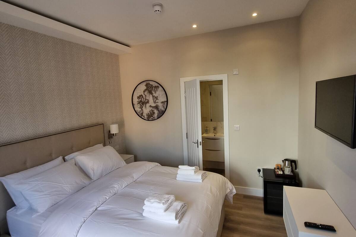 A modern hotel room with a large bed, a bedside table, a TV, and a bathroom visible through an open door.