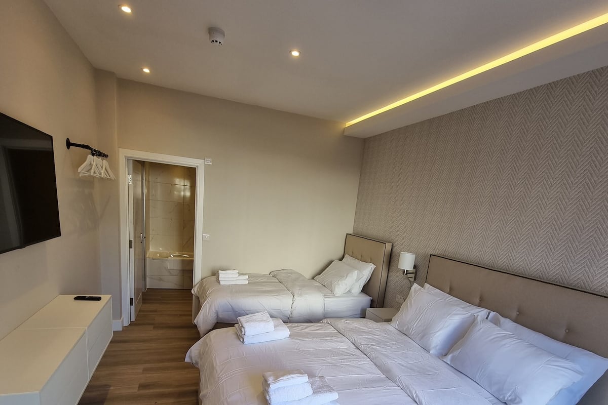 A modern hotel room with a large bed, a bedside table, a TV mounted on the wall, and a bathroom visible through an open door.