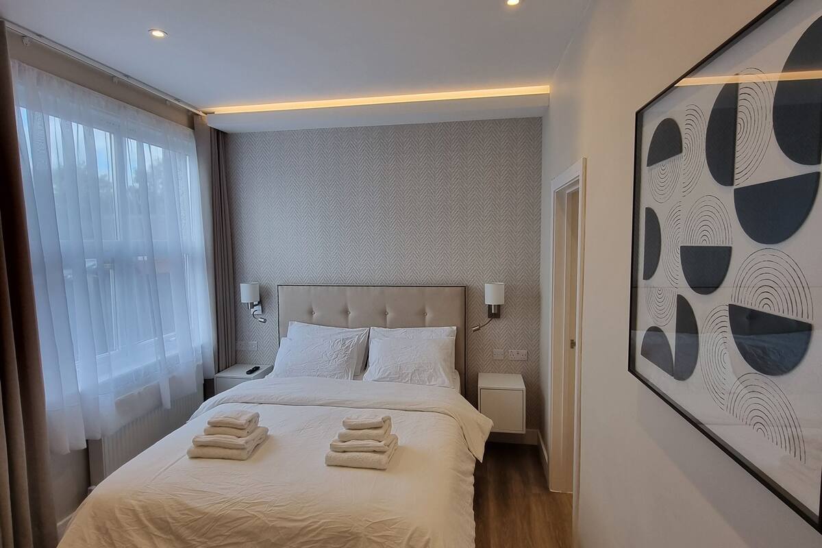 A modern hotel room with a large bed, a headboard, bedside tables, a wall-mounted light, and a framed abstract artwork.