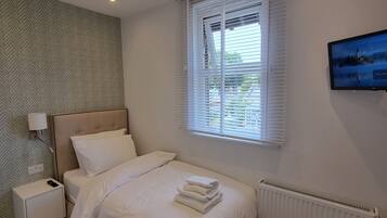 A bedroom with a bed, a nightstand, a wall-mounted TV, a window with blinds, and a radiator.