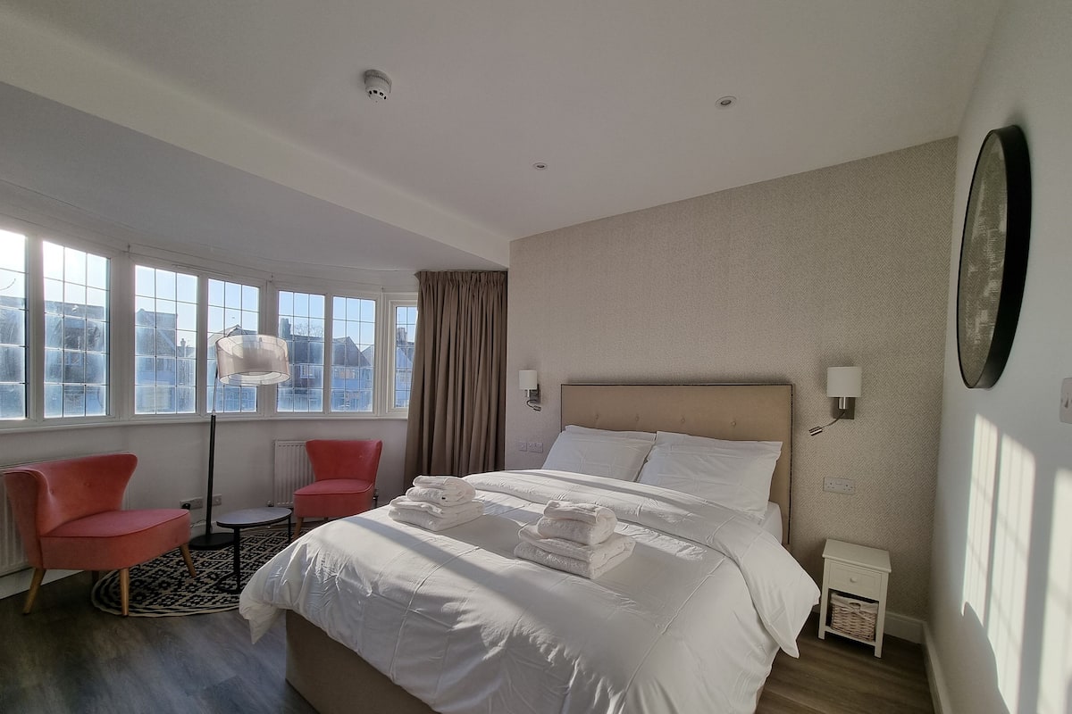 A modern hotel room with a large bed, two pink chairs, a floor lamp, and a view of buildings through large windows.