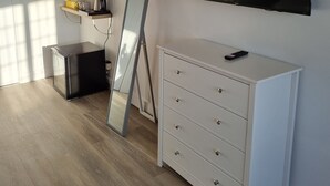 A modern room with a large flat-screen TV mounted on the wall, a white dresser with drawers, a small black cabinet, and a mirror leaning against the wall. - Hotel CG London (London)