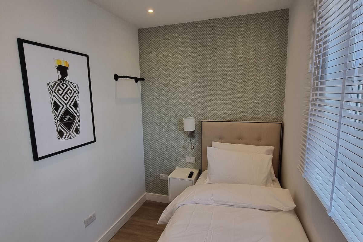 A hotel room with a bed, a nightstand, a wall-mounted lamp, a framed picture, and a window with blinds.