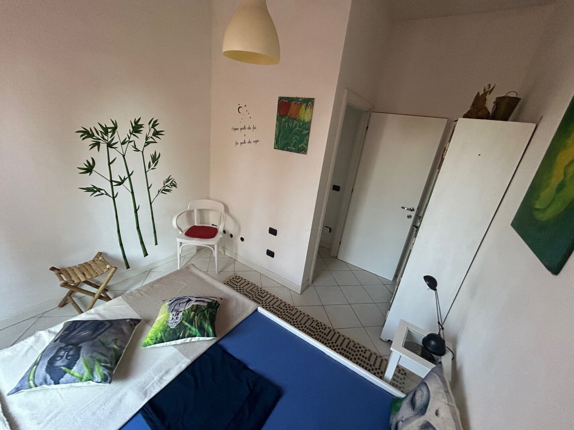 A bedroom with a bed, a chair, a wardrobe, and a wall with a bamboo plant and a painting.