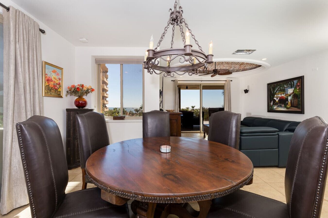 A dining area with a round wooden table surrounded by leather chairs, a chandelier, and a view through a glass door to an outdoor balcony.