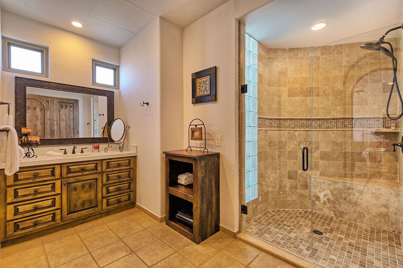 A bathroom with a wooden vanity, a large mirror, a shower with a glass door, and a tiled floor.