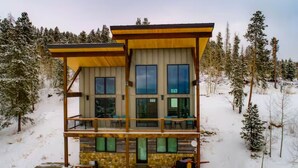 Une cabane à deux étages, avec une façade en bois et de grandes fenêtres, entourée d’arbres couverts de neige.