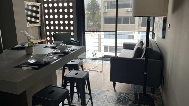 A modern kitchen with a central island, a dining area with a sofa, and a view of a building through large windows. - Increible depto en la Condesa cama King Size (Ciudad de México)