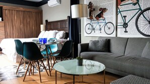 A modern hotel room with a grey sofa, a glass coffee table, and a dining area with blue chairs.
