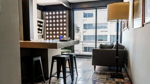 A compact living space with a kitchenette, a bar area, a sofa, and a large window offering a view of the city.