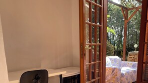 Una habitació amb escriptori, cadira, llit amb estores florals i vista a arbres a través d'una finestra de fusta.