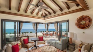 Un salón amplio con un gran ventanal que ofrece vistas al océano, un ventilador de techo y un espejo decorativo con diseño de sol.