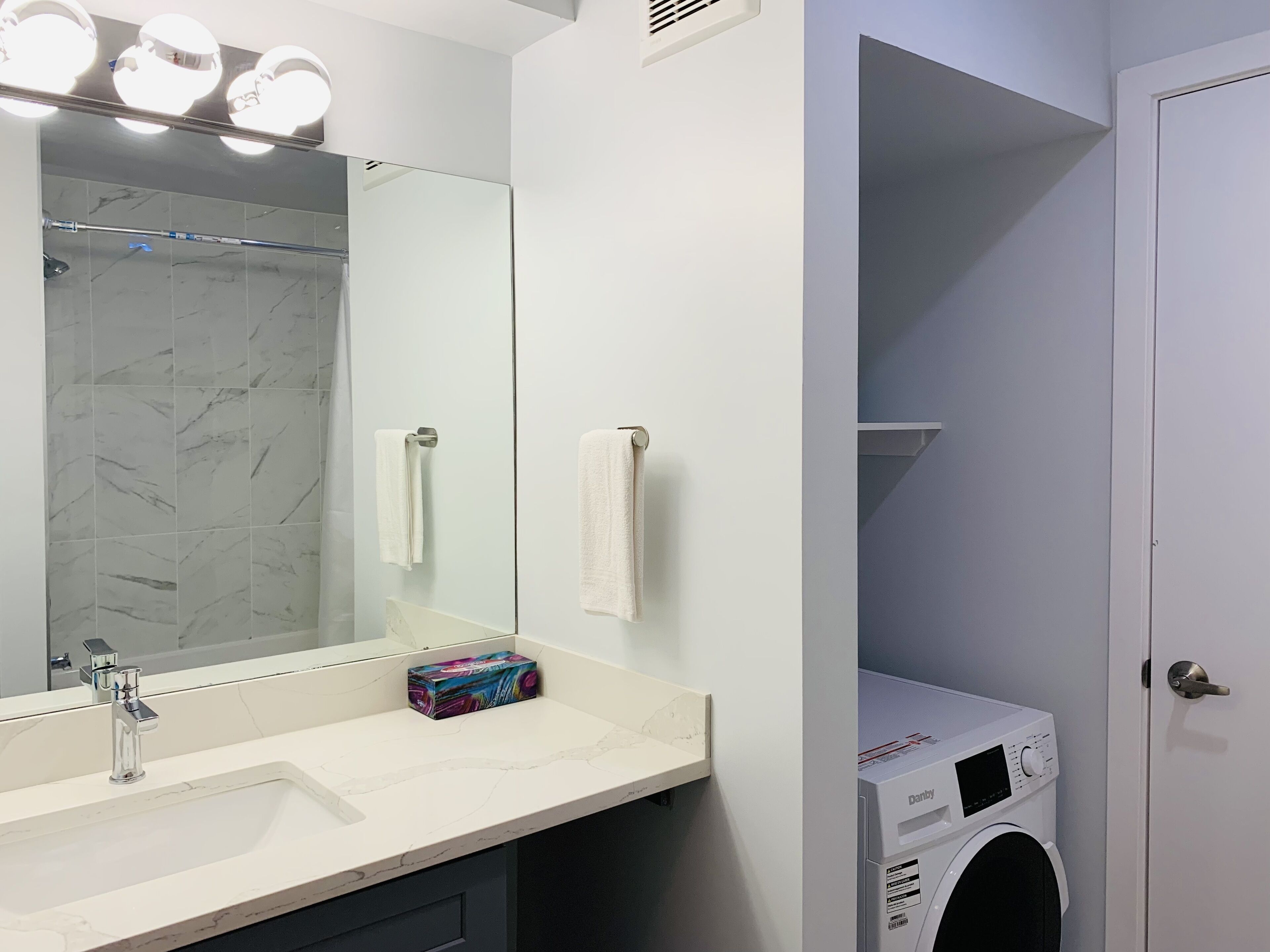 A bathroom with a marble sink, a large mirror, a towel rack, a washing machine, and a shelf.