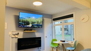 A modern room with a flat-screen TV mounted on the wall, a fireplace below, a dining area with a table and chairs, and a large window offering a view of the outdoors. - A Lovely Studio with Full Kitchen & AC (Canmore)