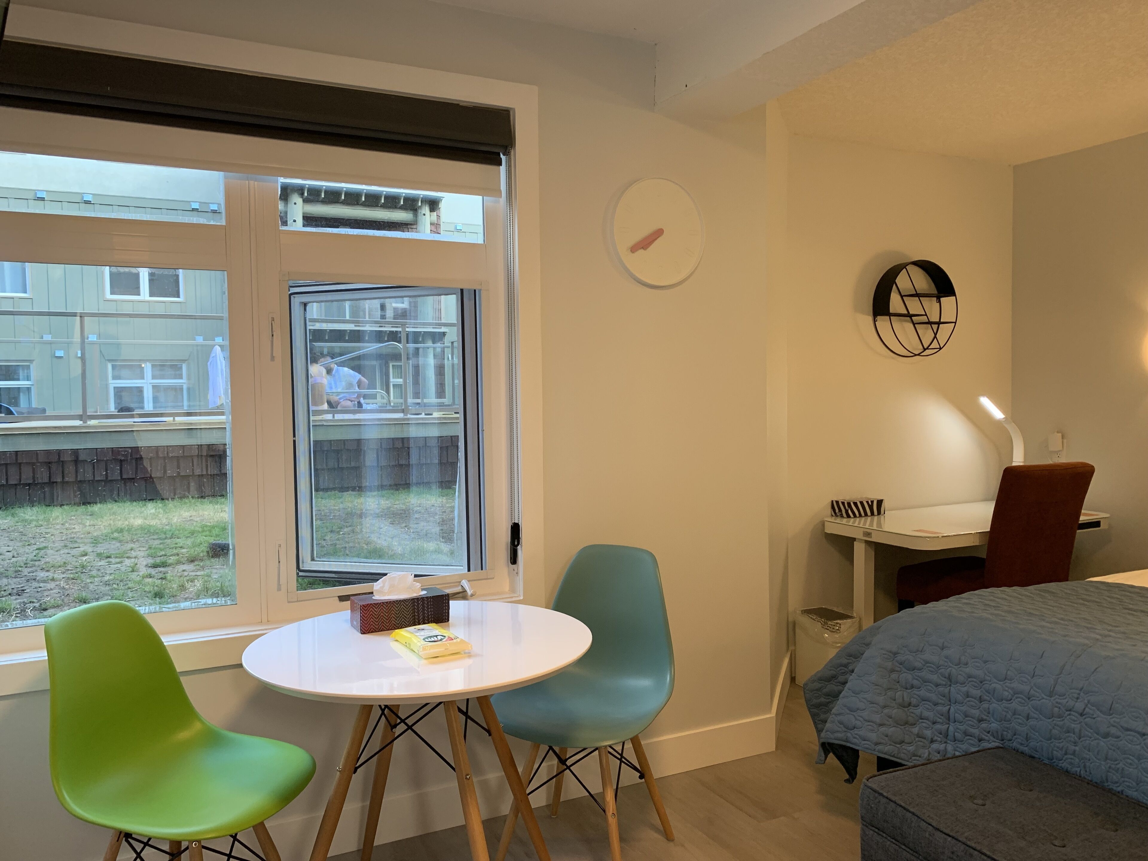 A small, modern room with a bed, a desk, and two chairs. There is a window with a view of a backyard and a building outside.