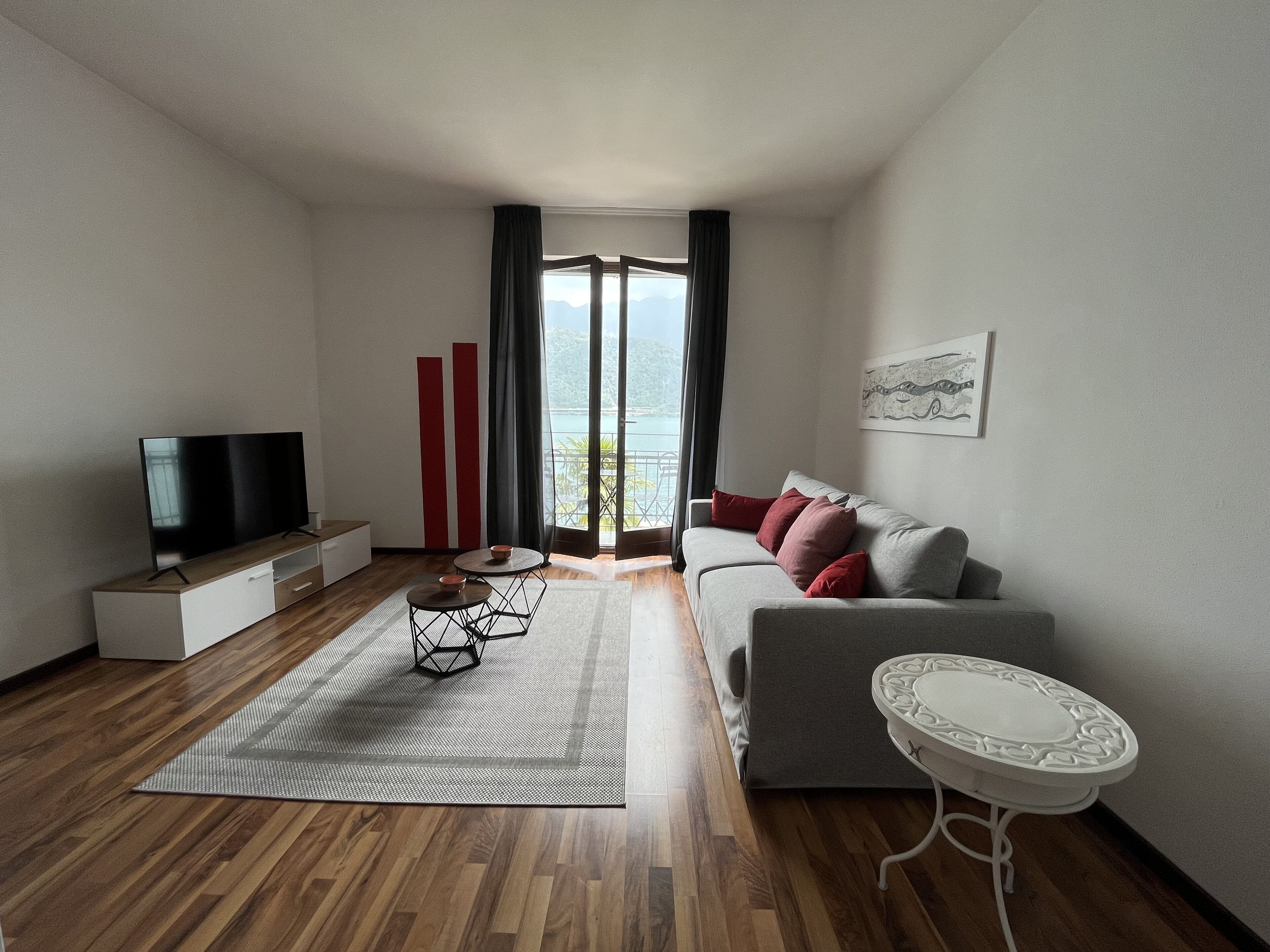 Una sala de estar moderna con un televisor de pantalla plana, un sofá gris con cojines rojos, una mesita auxiliar blanca y vistas al exterior a través de un ventanal grande.