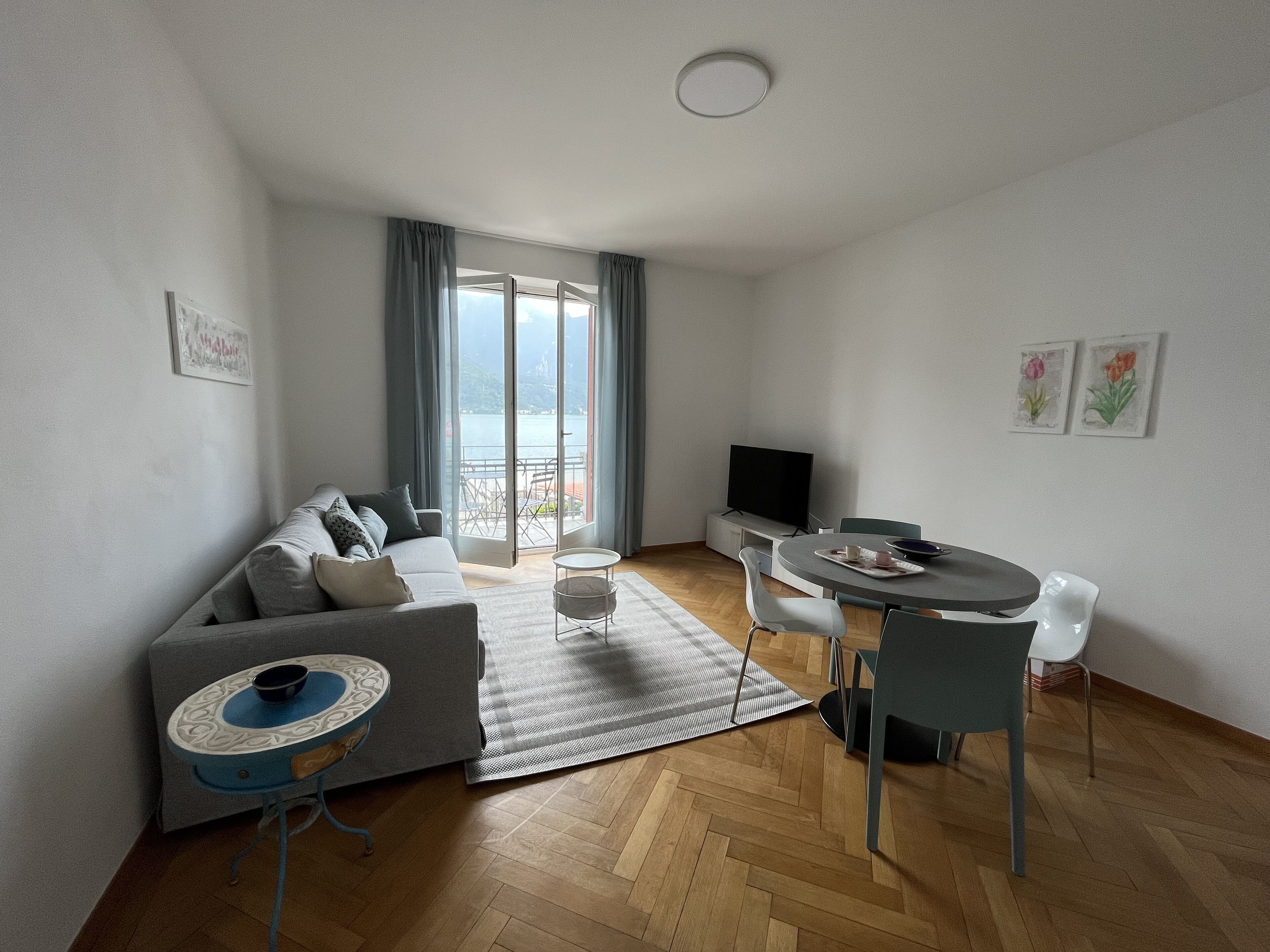 A modern living room with a sofa, a dining table with chairs, a TV, and a view of a body of water through large windows.