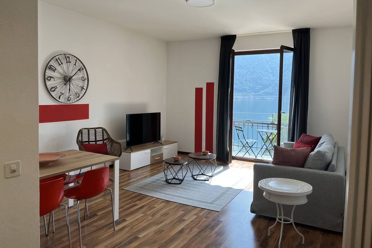 A modern living room with a flat-screen TV, a dining area with red chairs, a sofa with cushions, and a large window overlooking a body of water.