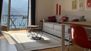 A modern living room with a sofa, red chair, and a table. A large window offers a view of mountains and a body of water. - 76thelakehouse (Melide)