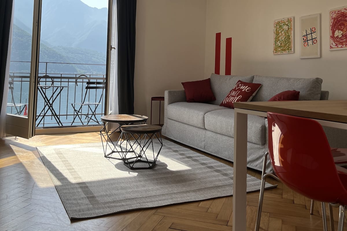 A modern living room with a sofa, red chair, and a table. A large window offers a view of mountains and a body of water.
