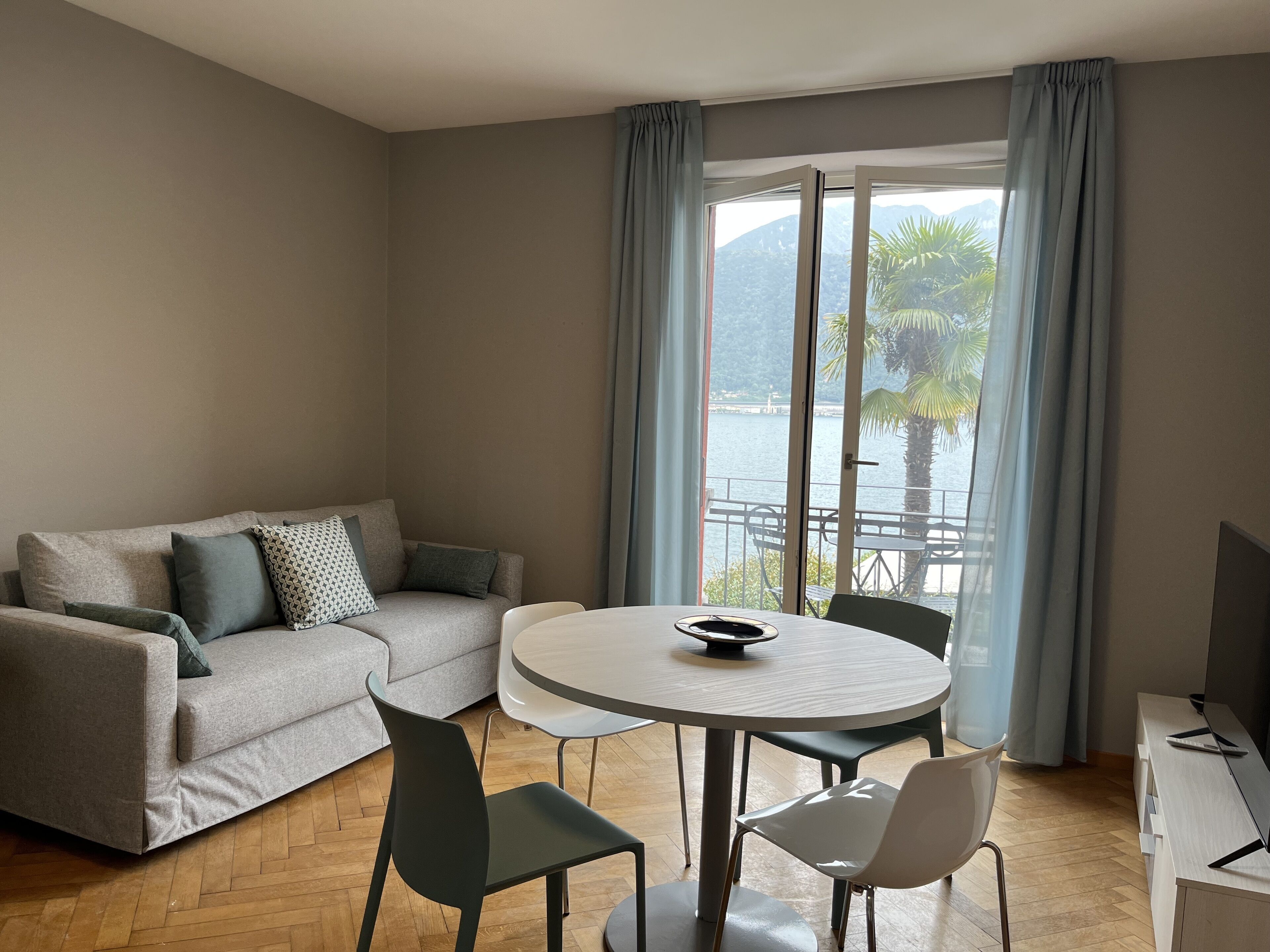 Ein modernes Wohnzimmer mit einem grauen Sofa, einem runden Esstisch und zwei Stühlen. Ein großes Fenster mit Vorhängen bietet Blick auf ein Gewässer und Palmen.