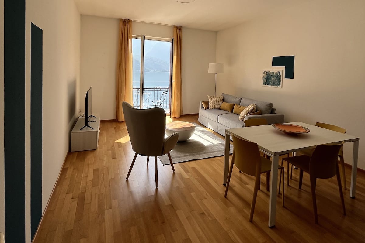 A modern living room with a dining area, wooden flooring, a sofa, a TV, and a balcony with curtains.