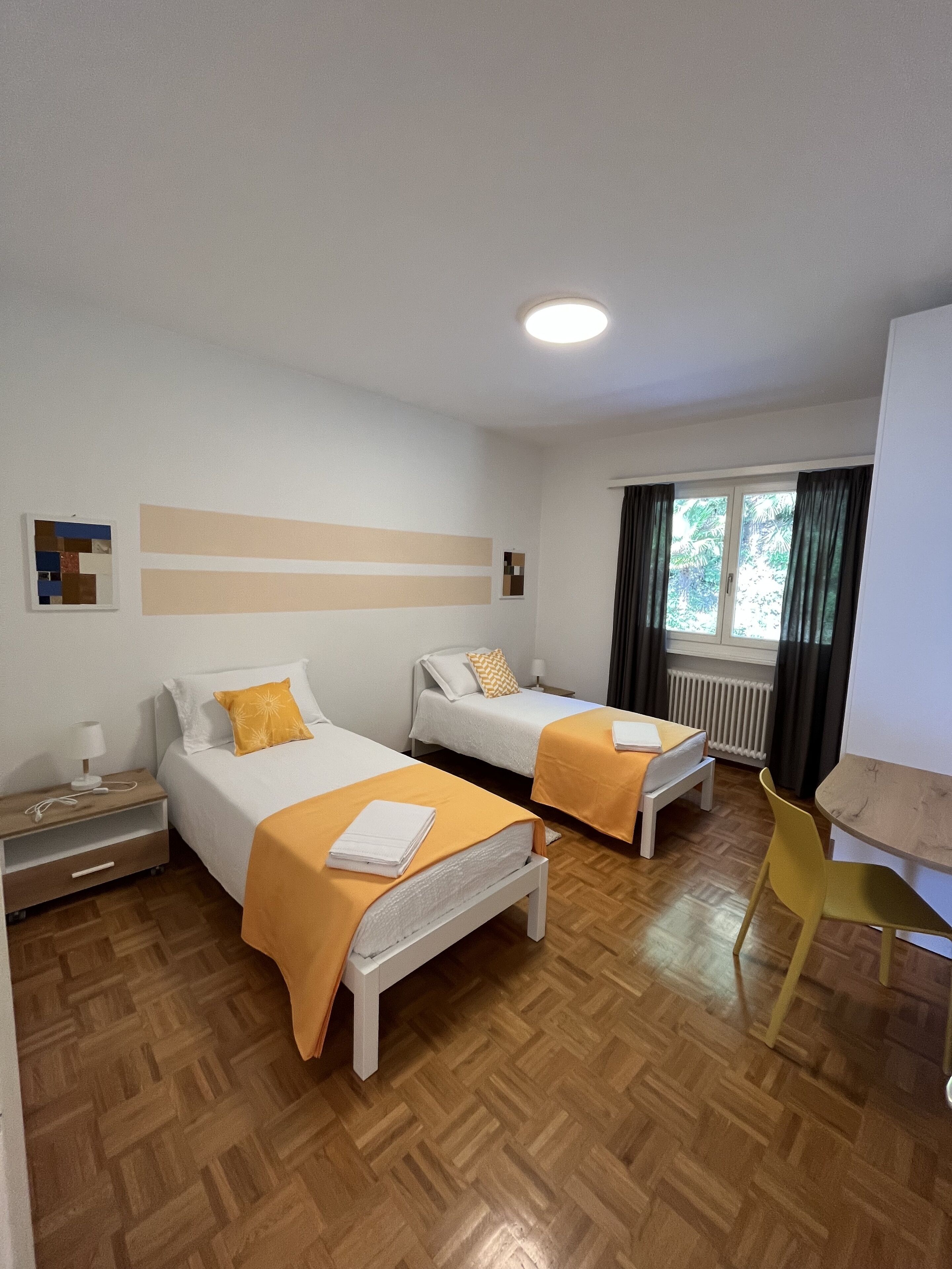 Ein Hotelzimmer mit zwei Betten, Holzboden, einem kleinen Tisch und einem Fenster mit Vorhängen.
