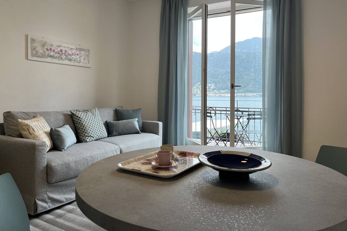 A modern living room with a grey sofa, a round table with a tray and cups, and a view of a lake and mountains through large windows.