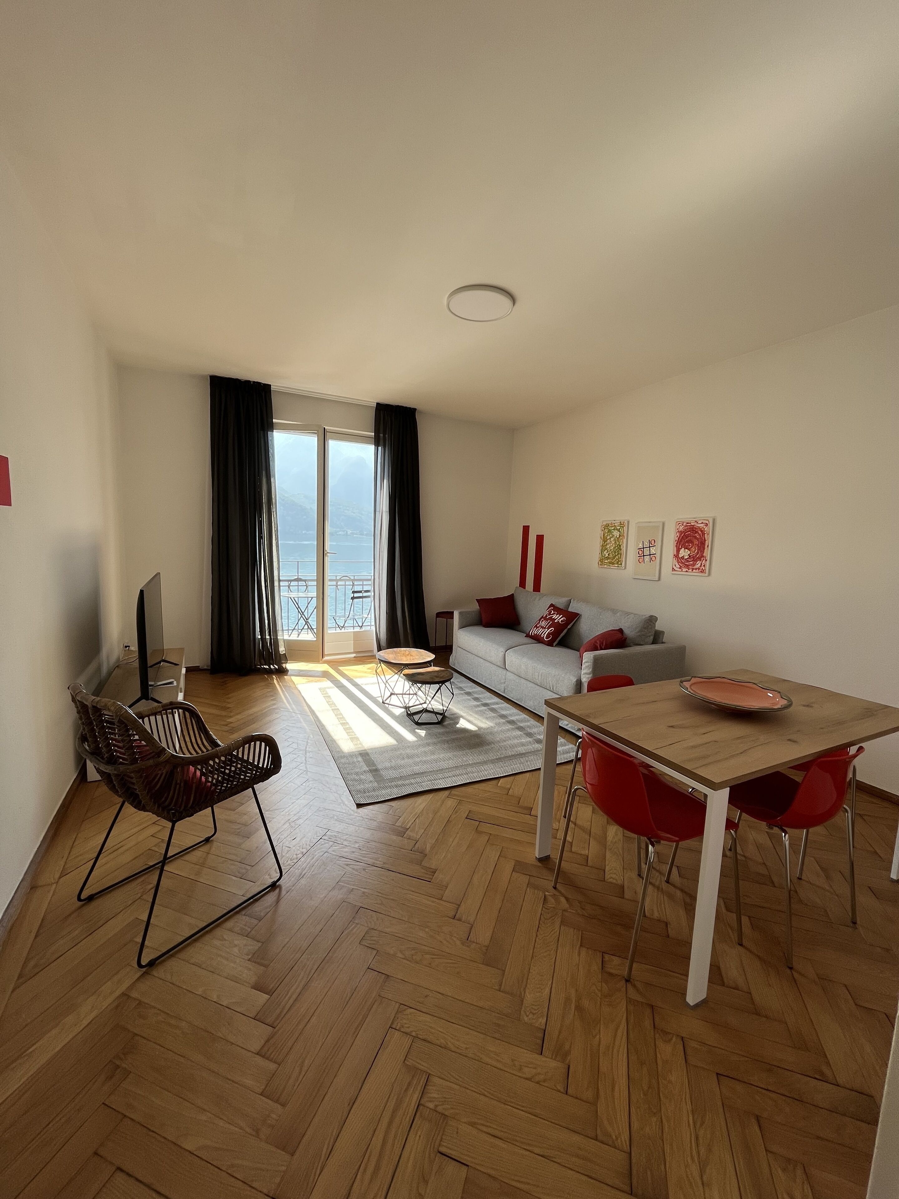 A modern living room with a wooden herringbone floor, a dining table with red chairs, a sofa with red cushions, and a large window with a view of the sea.