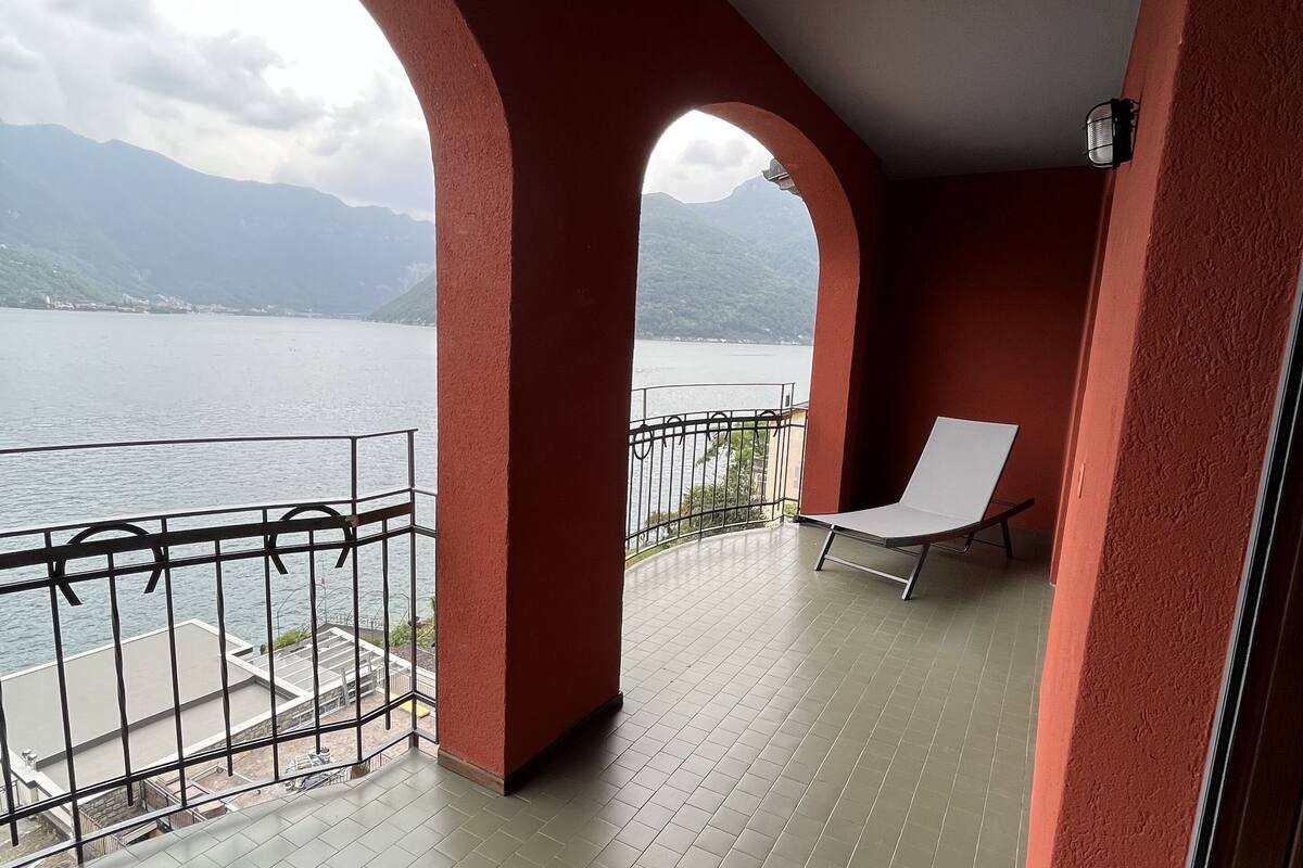 A balcony with a view of a lake and mountains, featuring a red wall, a white chair, and a tiled floor.