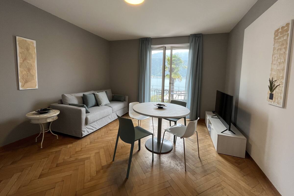 A modern living room with a grey sofa, a round dining table with chairs, a small side table, and a flat-screen TV.