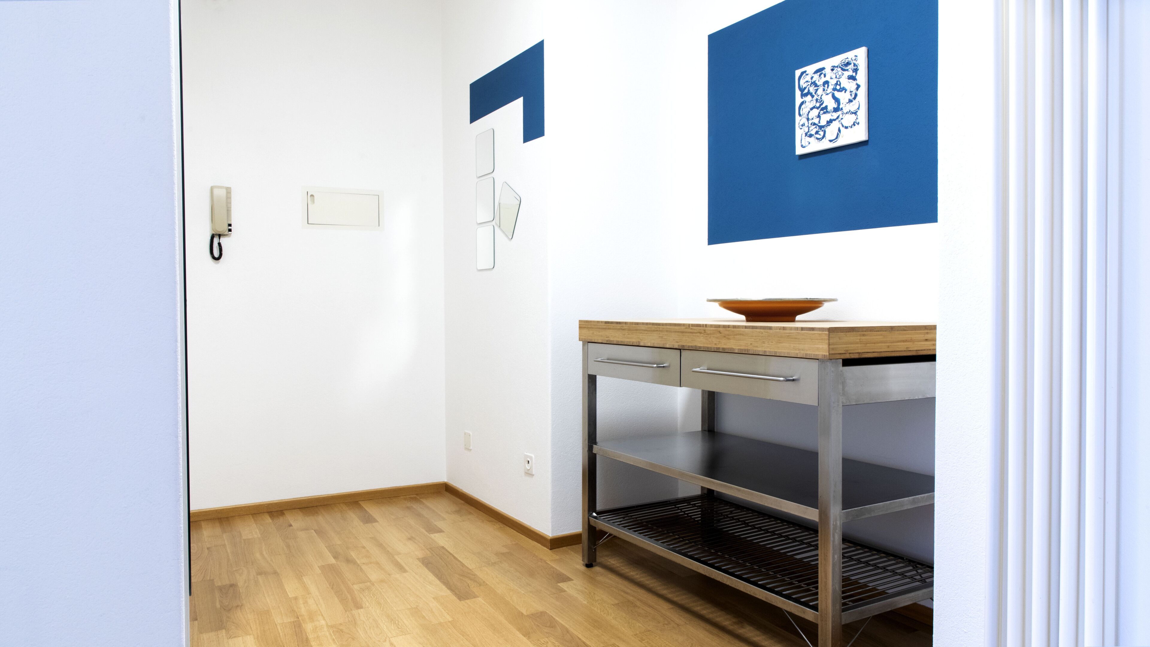 Ein Flur mit Holzboden, einem Tisch aus Edelstahl mit einer Schüssel und einer blauen Wand mit weißem Muster.
