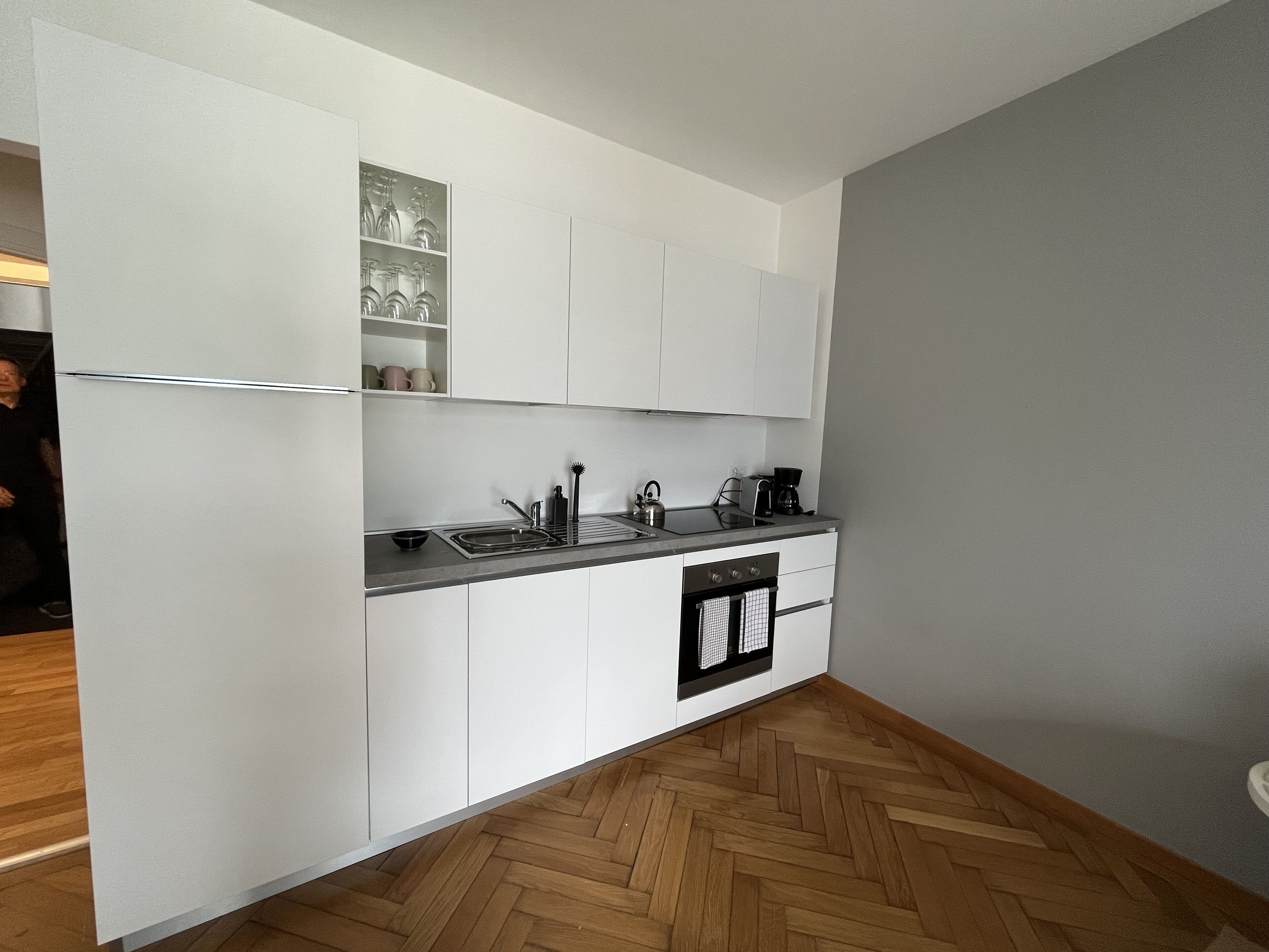 A compact kitchen with white cabinetry, a herringbone-patterned wooden floor, and a built-in oven.